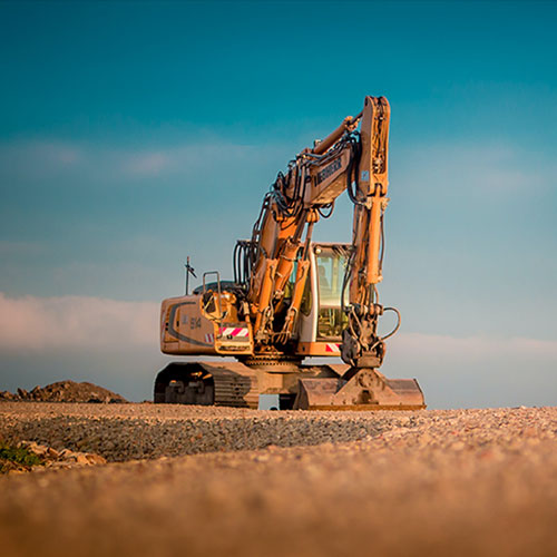 A single excavator parked on a gravel construction site under a clear blue sky, with the machine facing forward and its arm resting on the ground.
