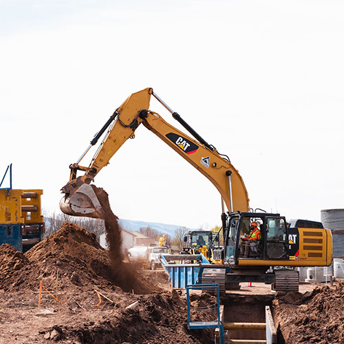 Construction site with a CAT excavator lifting a bucket of dirt over a trench, operated by a worker in safety gear, with equipment and soil piles in the background.