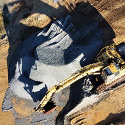 Aerial view of an excavator scooping gravel from a circular excavation area surrounded by dirt, with sharp shadows cast by the machine.
