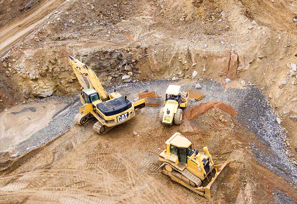 Aerial view of a construction site with three large yellow machines—an excavator, a wheel loader, and a bulldozer—working on a rocky, dirt-filled terrain. The area appears to be an excavation zone with exposed layers of earth and stone.